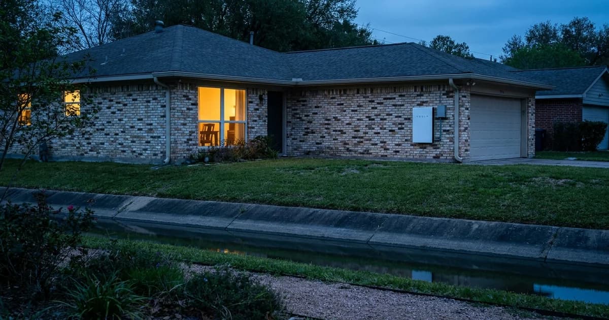 A single-story brick ranch home in north Houston at blue hour, a white wall-mounted home battery unit visible on the garage wall at chest height, warm amber light from the window, a drainage bayou channel with grassy banks in the background.