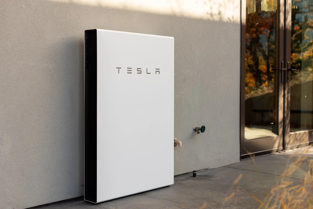 Tesla Powerwall 3 home battery unit leaning against an exterior wall, showing the clean white form factor and TESLA branding
